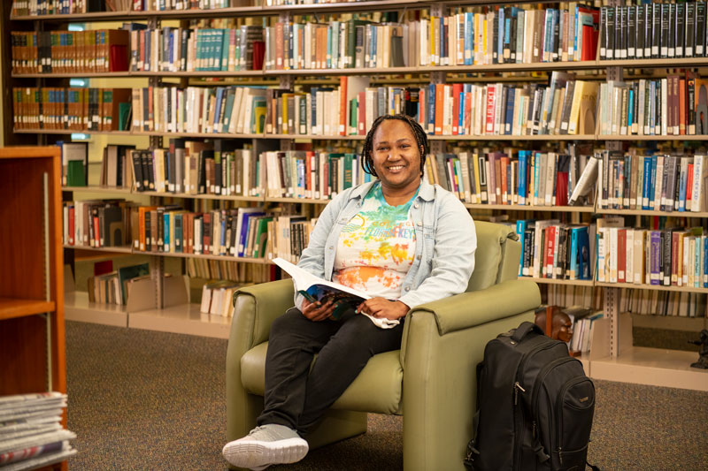 Black female college student sitting in a chair smiling, in a library, in front of a shelf of books. She's holding a book and her backpack is on the floor next to her.