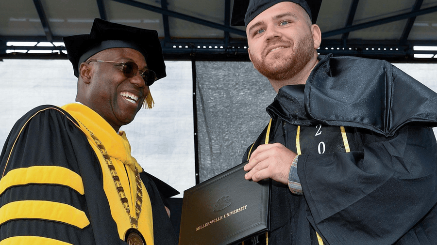 A Black man with a big smile wearing black and gold robes confers a degree to a white male student. The student is facing the audience smiling, holding his diploma. 