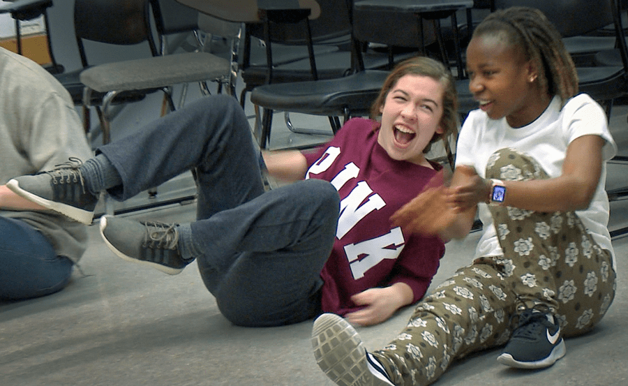 Two girls laughing together on the floor in dance class. They are dressed casually, in sweats, one is leaning back on her arm with her feet off the ground, the other has one leg straight in front of her and the other bent with her arm around it.